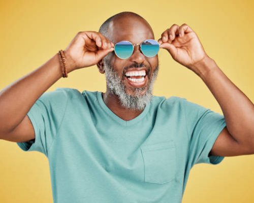 Happy mature African American man standing alone against yellow background in a studio and posing with sunglasses. Smiling black man feeling fashionable and cool while wearing glasses. Summer and beach